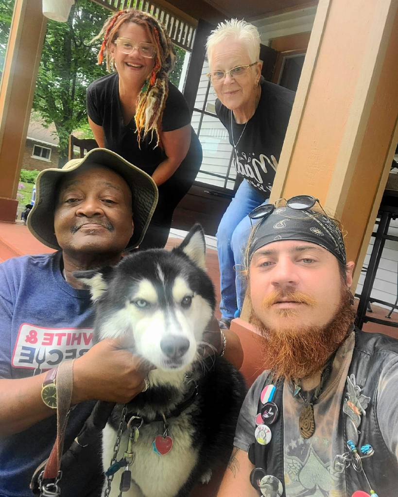 A group portrait featuring four people and a black and white husky dog. Two women are standing in the background, one with colorful dreadlocks and the other with short gray hair and glasses. A man sits in the foreground holding the dog, while another man takes a selfie. The setting appears to be a porch with a house visible in the background.